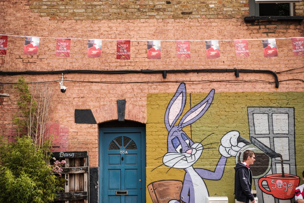 A mural around a front door.  It features cartoon character Buggs Bunny pouring coffee into a red cup.  In the foreground a man walks by wearing a red and white scarf and above his head is bunting with the words "lovely day to be champions"