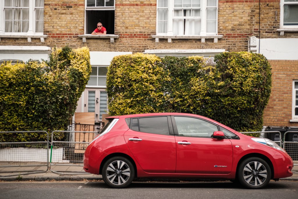 A man in a red shirt at the open window of a house.  In the foreground is a red car.