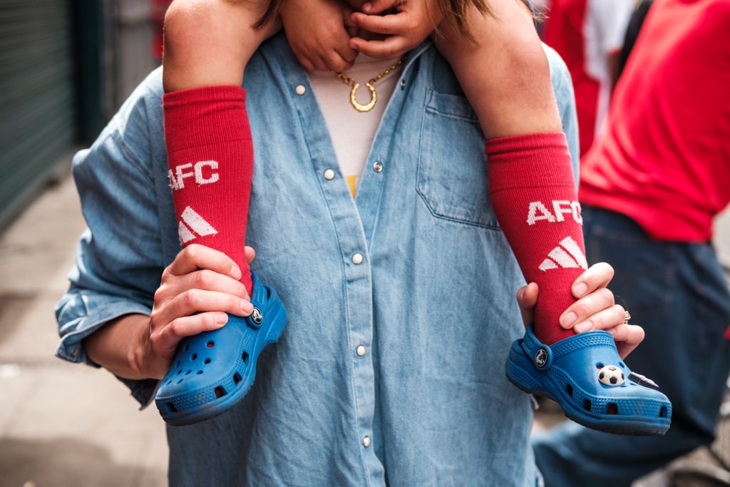 A child sat on a woman's shoulders.  We see his legs from the knees down and he is wearing red socks with "AFC" embroidered on the front.  He is also wearing blue crocks shoes.