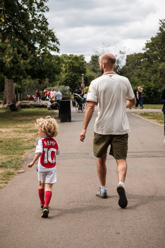 A man and a small boy walk through the park.  The boy is wearing a football shirt with "REMY" and the number ten on the back.