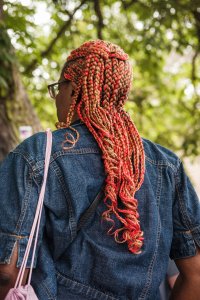 A woman of the African diaspora has her back to the camera.  She is wearing a denim jacket against which lie her braided hair plaited with the colours of yellow and red