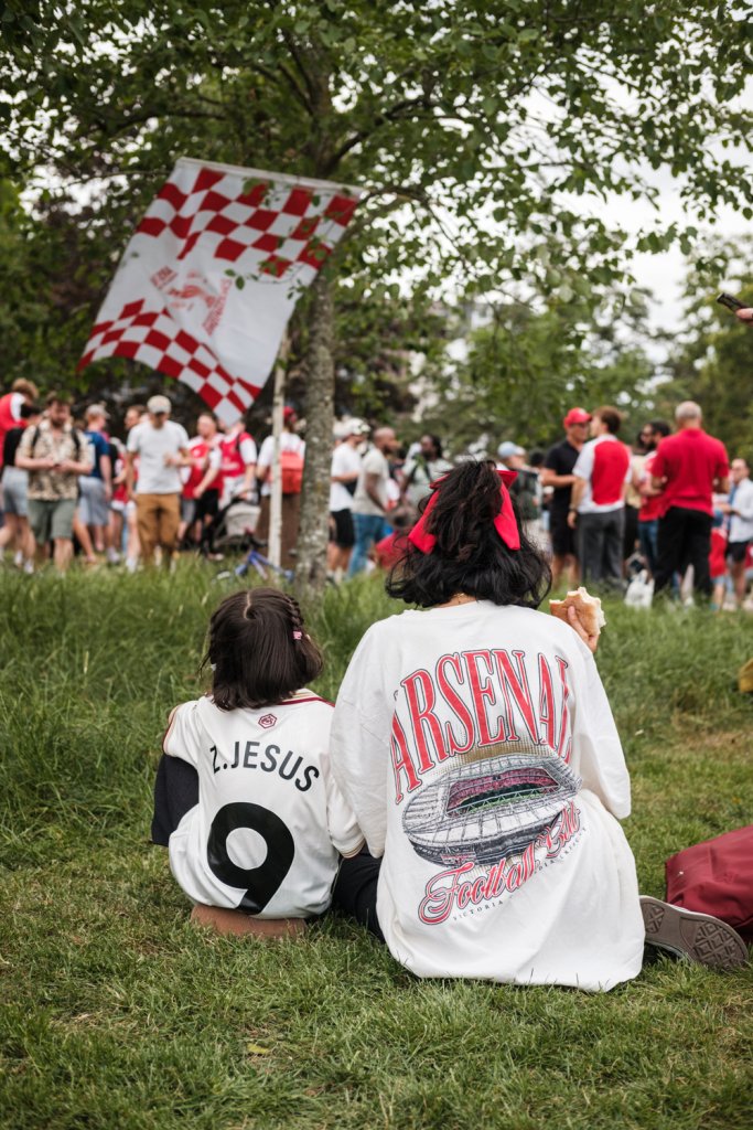 In the background are gathered a group of football supporters.  In the foreground a woman sits with a child.  The child wears a number nine shirt with "z.jesus"