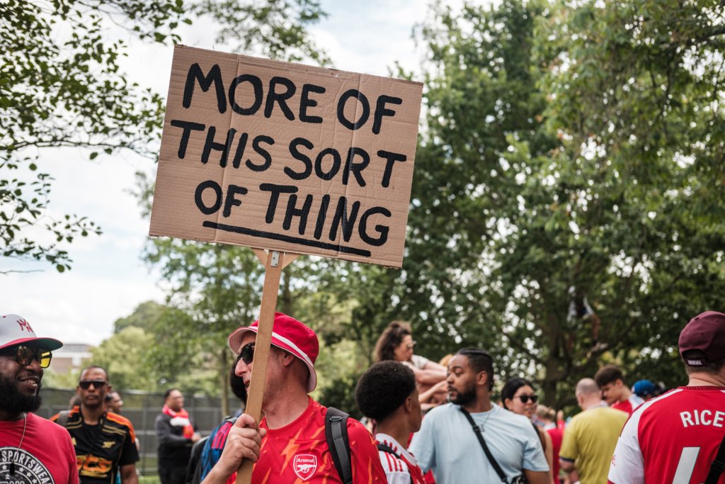 Set in a park a group of people in the background.  In the foreground a man is holding a homemade sign with the words "more of this sort of thing" He is wearing an Arsenal football shirt.